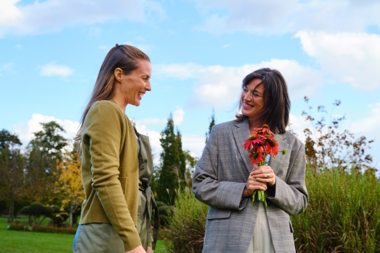 Zwei Frauen stehen in einem Garten, eine von ihnen hält einen Strauß roter Blumen