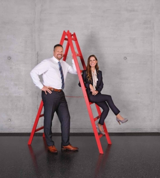 Lächelnde Frau sitzt auf roter Leiter. Ein lächelnder Mann steht neben ihr.