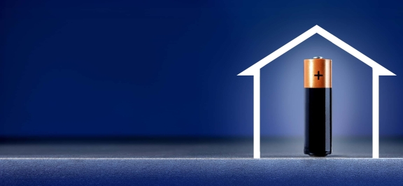 Jetzt Wohnen Artikel: Stromspeicher für zu Hause - blauer Hintergrund mit weiße Umrandung eines Hauses, mit Batterie innerhalb des Hauses