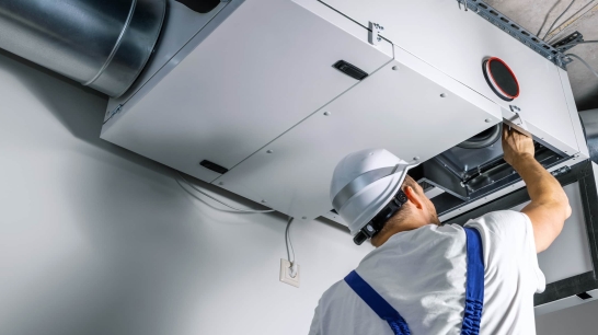 Jetzt Wohnen Artikel: Wohnraumlüftung für zuhause. Elektriker:in arbeitet an einer Wohnraumlüftung an der Decke eines Raumes.